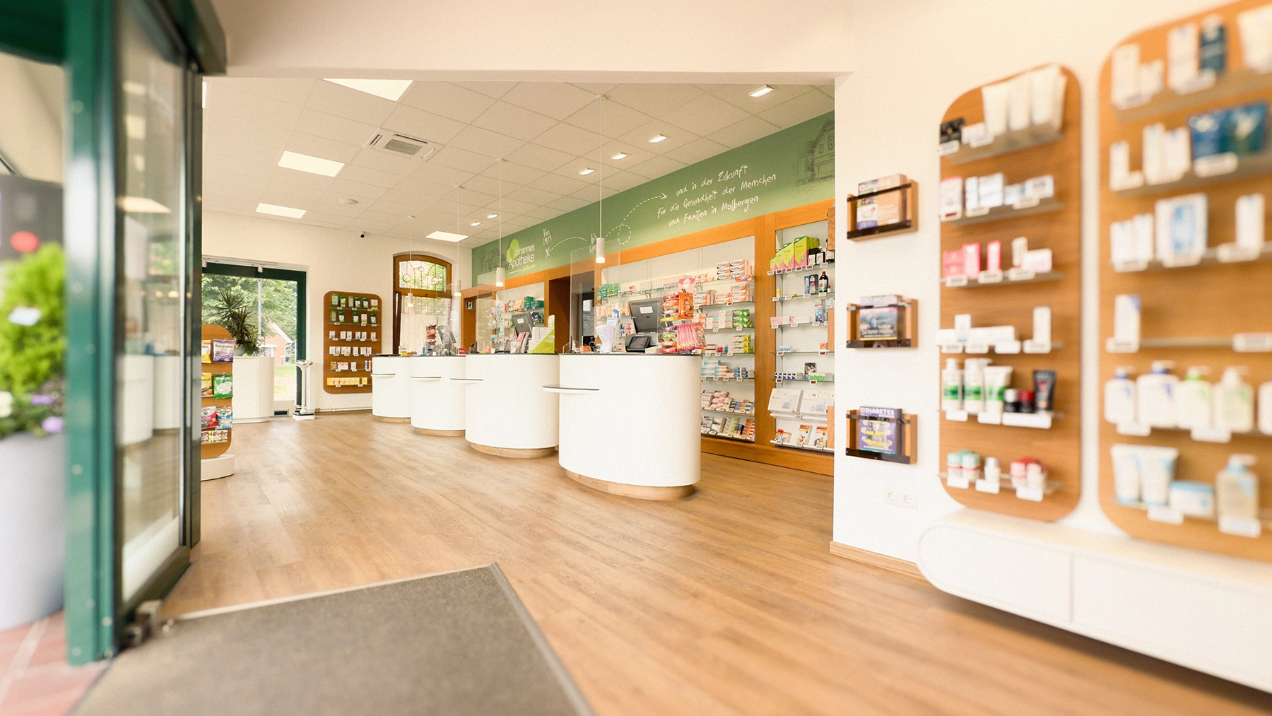 Blick durch den Eingang in die Johannes Apotheke Molbergen mit hellen, modernen Beratungstresen, Holzfußboden und übersichtlich angeordneten Regalen mit Gesundheits- und Pflegeprodukten.